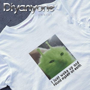 May include: White t-shirt featuring a green alien-like creature with the text "I just woke up and found myself on earth." The design is printed on the front of the shirt. The shirt has a crew neck.