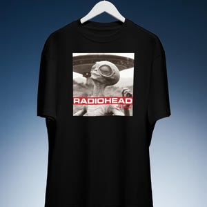 May include: Black t-shirt featuring a graphic of an alien with a UFO in the background. The text "RADIOHEAD" is displayed in red above the alien image, with "the bends" below. The shirt is hanging on a white hanger.