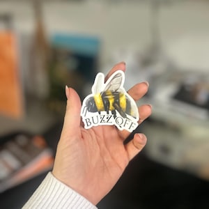 Puede incluir: Una pegatina blanca con una ilustración realista de un abejorro con rayas negras y amarillas. La pegatina tiene el texto "Buzz Off" en letras negras.
