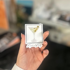 May include: A white sticker with a black border and a drawing of a martini glass with olives. The text 