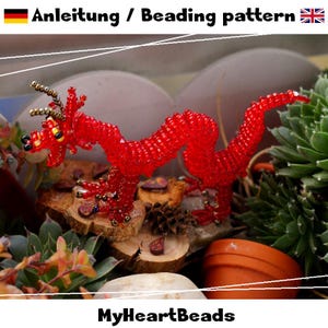 Może przedstawiać: Figurka smoka wykonana z jaskrawoczerwonych koralików z metalicznymi złotymi i czarnymi akcentami na rogach i oczach. Tekst "Anleitung / Beading pattern" oraz "MyHeartBeads" jest widoczny u góry i u dołu obrazu.