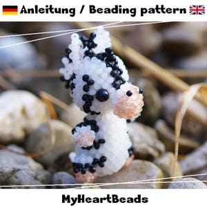 May include: A beaded zebra figurine, crafted with white and black beads, and a pinkish-beige snout and feet. The text "Anleitung / Beading pattern" is at the top, and "MyHeartBeads" is at the bottom. The zebra is set against a background of rocks and dried grass.
