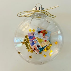 May include: A clear glass Christmas ornament filled with gold star-shaped confetti and small, colorful book illustrations. The ornament is topped with a silver cap and a gold ribbon bow. The illustrations appear to be from a children's book.
