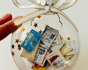 May include: A clear spherical glass bauble filled with miniature book replicas by Robin Hobb, including titles like Ship of Magic and Fool's Quest. The bauble features gold star confetti inside and is topped with a white ribbon with gold edges.