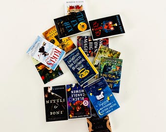 May include: A collection of miniature books with colorful covers and titles like "The Hex," "Queen B," and "Nettle & Bone." The books are arranged on a white surface, showcasing various fantasy and witch-themed titles.