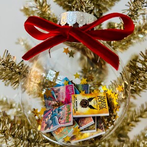 May include: A clear spherical ornament filled with miniature Harry Potter book replicas and gold star confetti. The ornament is topped with a silver cap and a vibrant red velvet ribbon bow, creating a festive display of literary-themed decor.