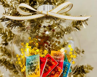 May include: A clear spherical Christmas ornament filled with gold star-shaped confetti and miniature book replicas. The books feature titles including A Court of Thorns and Roses and A Court of Silver Flames. A white ribbon with gold trim is tied at the top.