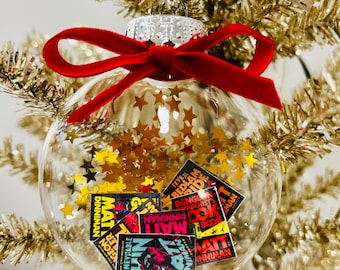May include: A clear spherical glass ornament filled with gold star-shaped confetti and miniature book covers featuring the title Dungeon Crawler Carl by Matt Dinniman. The ornament is topped with a silver cap and a bright red velvet ribbon bow.
