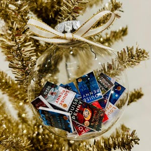 May include: A clear glass spherical ornament filled with miniature book replicas by author Louise Penny. The ornament is topped with a white ribbon featuring gold trim. The visible book titles include Glass Houses, A World of Curiosities, and Bury Your Dead.