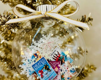 May include: A clear spherical Christmas ornament filled with white plastic snowflakes and miniature book covers, including titles like Heated Rivalry by Rachel Reid. The ornament is topped with a white ribbon featuring gold trim.