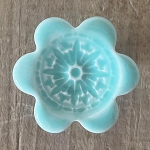 May include: A light blue ceramic flower-shaped dish with a detailed central design. The dish has six rounded petals and a glossy finish. The design in the center is a detailed pattern. The dish is on a wooden surface.