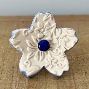 May include: A flower-shaped ceramic knob with a cream-colored surface and blue edges. The knob features a floral pattern and a dark blue center. This decorative hardware is suitable for cabinets and drawers.