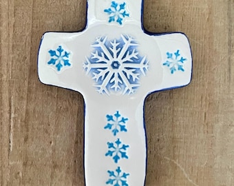 Op de afbeelding: Witte keramische kruisornament met blauwe sneeuwvlokpatronen en een blauwe rand. Het ornament hangt aan een zilveren lint. Het kruis is versierd met een grote sneeuwvlok in het midden en kleinere op de armen.