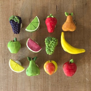 Pu&ograve; includere: Una collezione di calamite colorate in ceramica a forma di frutta, tra cui uva, lime, fragola, pera, mela, anguria, uva, banana, limone, pera, mela e mela rossa. Le calamite sono disposte su una superficie di legno.