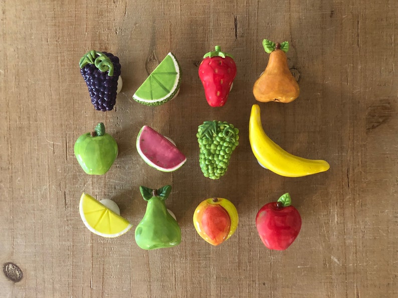 Pu&ograve; includere: Una collezione di calamite colorate in ceramica a forma di frutta, tra cui uva, lime, fragola, pera, mela, anguria, uva, banana, limone, pera, mela e mela rossa. Le calamite sono disposte su una superficie di legno.