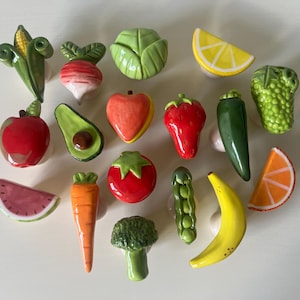Pu&ograve; includere: Una collezione di quindici pomelli in ceramica per mobili a forma di vari frutti e verdure, tra cui una pannocchia, un ravanello, un cavolo, una fetta di limone, una mela, un avocado, una pesca, una fragola, un peperoncino, uva, anguria, carota, pomodoro, broccoli, piselli, banana e una fetta d'arancia.