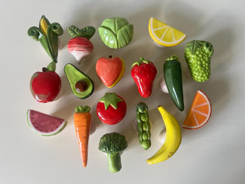 Pu&ograve; includere: Una collezione di quindici pomelli in ceramica per mobili a forma di vari frutti e verdure, tra cui una pannocchia, un ravanello, un cavolo, una fetta di limone, una mela, un avocado, una pesca, una fragola, un peperoncino, uva, anguria, carota, pomodoro, broccoli, piselli, banana e una fetta d'arancia.