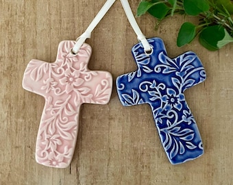 May include: Two ceramic cross ornaments. One is light pink with a floral design, and the other is blue with a white floral pattern. Each cross has a white ribbon for hanging. The crosses are displayed on a wooden surface.