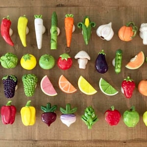 Pu&ograve; includere: Una collezione di pomelli per mobili in ceramica colorata a forma di verdura e frutta. I pomelli includono un avocado, peperoni, carote, uno spicchio d'aglio, una zucca, una banana e altro ancora. Sono disposti su una superficie di legno.