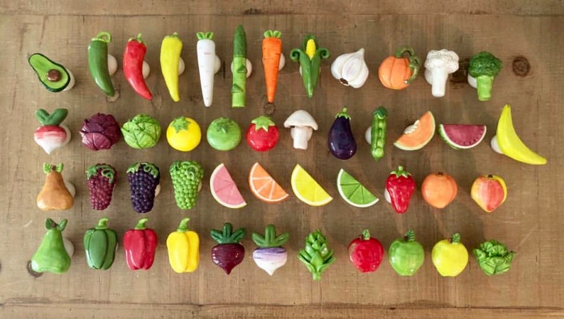 Pu&ograve; includere: Una collezione di pomelli per mobili in ceramica colorata a forma di verdura e frutta. I pomelli includono un avocado, peperoni, carote, uno spicchio d'aglio, una zucca, una banana e altro ancora. Sono disposti su una superficie di legno.