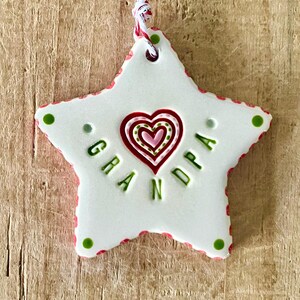 Puede incluir: Adorno blanco en forma de estrella con la palabra "GRANDPA" en letras verdes. El adorno presenta un dise&ntilde;o de coraz&oacute;n en rojo y rosa, y est&aacute; adornado con detalles rojos y verdes. Se adjunta una cuerda roja y blanca para colgar.