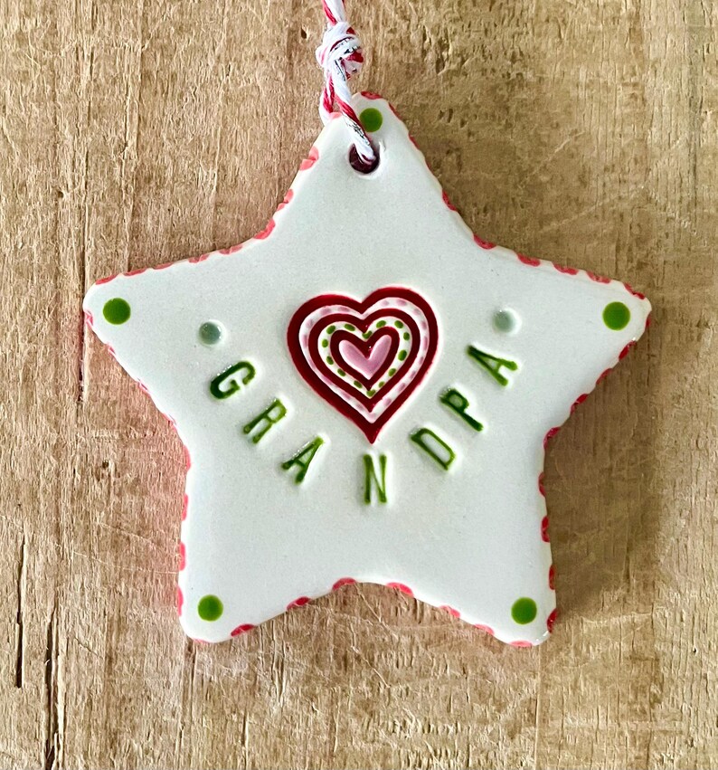Puede incluir: Adorno blanco en forma de estrella con la palabra "GRANDPA" en letras verdes. El adorno presenta un dise&ntilde;o de coraz&oacute;n en rojo y rosa, y est&aacute; adornado con detalles rojos y verdes. Se adjunta una cuerda roja y blanca para colgar.
