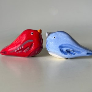 Peut inclure: Deux figurines d'oiseaux en céramique, l'une rouge et l'autre bleue, se faisant face. L'oiseau rouge a un bec jaune et des accents blancs. L'oiseau bleu a un ventre blanc et des accents blancs.