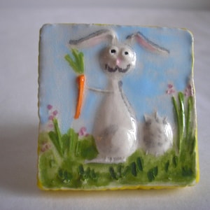 Op de afbeelding: Een keramische tegel met een cartoonkonijn dat een wortel vasthoudt. Het konijn is wit met grijs haar en heeft een blauwe achtergrond met groen gras en roze bloemen.
