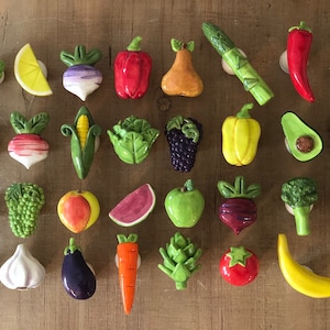 Pu&ograve; includere: Una collezione di calamite in ceramica a forma di frutta e verdura, tra cui un peperone rosso, asparagi verdi e una banana gialla. Le calamite sono disposte su una superficie di legno.