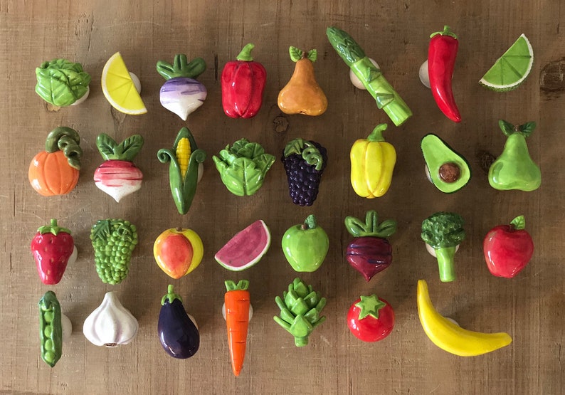 Pu&ograve; includere: Una collezione di calamite in ceramica a forma di frutta e verdura, tra cui un peperone rosso, asparagi verdi e una banana gialla. Le calamite sono disposte su una superficie di legno.