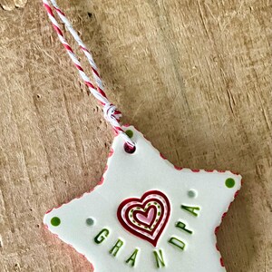 Puede incluir: Un adorno blanco en forma de estrella con la palabra "GRANDPA" en letras verdes. Un dise&ntilde;o de coraz&oacute;n rojo y rosa est&aacute; en el centro. El adorno tiene un borde rojo y verde y una cuerda roja, blanca y plateada para colgar.