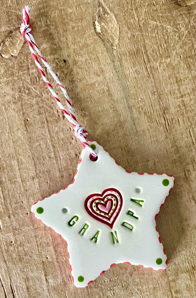 Puede incluir: Un adorno blanco en forma de estrella con la palabra "GRANDPA" en letras verdes. Un dise&ntilde;o de coraz&oacute;n rojo y rosa est&aacute; en el centro. El adorno tiene un borde rojo y verde y una cuerda roja, blanca y plateada para colgar.