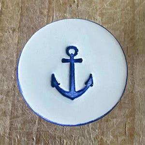 May include: A white ceramic coaster featuring a blue anchor design. The anchor is centrally positioned on the coaster and has a simple, classic style.