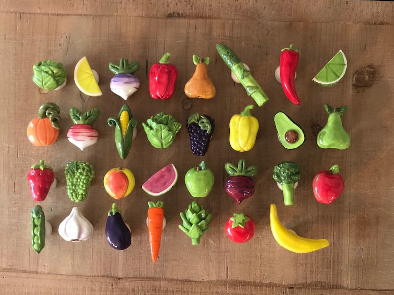 Pu&ograve; includere: Una collezione di calamite in ceramica colorate raffiguranti verdure e frutta, tra cui un peperone rosso, una banana gialla e un avocado verde. Le calamite sono disposte su una superficie di legno.