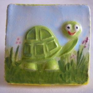 Könnte beinhalten: Eine handbemalte Keramikfliese mit einer grünen Schildkröte mit einem lächelnden Gesicht vor einem blauen Himmel. Die Schildkröte hat einen detaillierten Panzer und ist von grünem Gras und rosa Blumen umgeben.