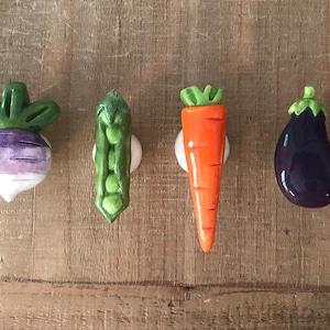 Pu&ograve; includere: Una collezione di pomelli per mobili in ceramica a forma di verdura. Il set comprende un broccolo verde, una rapa viola e bianca, un baccello di piselli verde, una carota arancione, una melanzana viola scuro e un pomodoro rosso. Ogni pomello &egrave; montato su una base bianca.