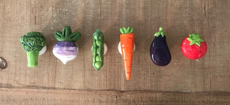 Pu&ograve; includere: Una collezione di pomelli per mobili in ceramica a forma di verdura. Il set comprende un broccolo verde, una rapa viola e bianca, un baccello di piselli verde, una carota arancione, una melanzana viola scuro e un pomodoro rosso. Ogni pomello &egrave; montato su una base bianca.