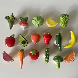 Pu&ograve; includere: Una collezione di quindici pomelli in ceramica per mobili a forma di vari frutti e verdure, tra cui mais, ravanello, cavolo, limone, uva, mela, avocado, pesca, fragola, peperoncino, banana, anguria, carota, pomodoro, pisello, broccoli e arancia.