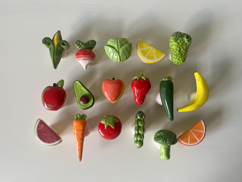 Pu&ograve; includere: Una collezione di quindici pomelli in ceramica per mobili a forma di vari frutti e verdure, tra cui mais, ravanello, cavolo, limone, uva, mela, avocado, pesca, fragola, peperoncino, banana, anguria, carota, pomodoro, pisello, broccoli e arancia.