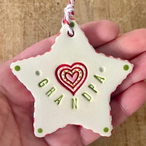 Puede incluir: Adorno de cer&aacute;mica en forma de estrella con la palabra "GRANDPA" en letras verdes. El adorno tiene un dise&ntilde;o de coraz&oacute;n rojo y rosa y un borde rojo. Se sujeta con una cuerda roja y blanca.