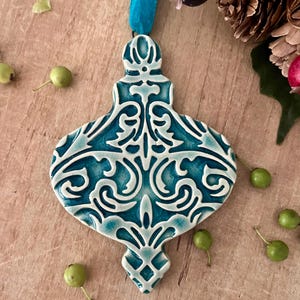 May include: A teardrop-shaped ceramic ornament featuring a glossy teal glaze with a white raised relief pattern of intricate scrolls and filigree. The ornament is suspended by a bright blue velvet ribbon attached to the top loop.