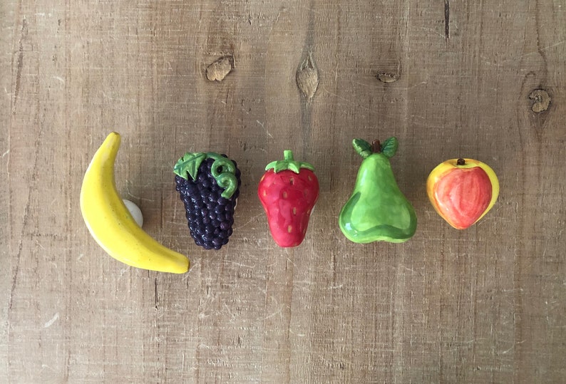 Pu&ograve; includere: Una collezione di calamite a forma di frutta in ceramica, tra cui una banana gialla, un grappolo d'uva viola, una fragola rossa, una pera verde e una mela rossa e gialla. Le calamite sono disposte su una superficie di legno.