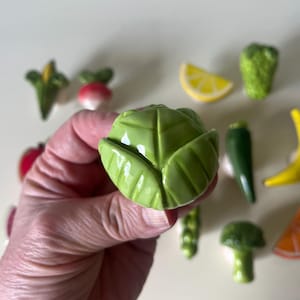 Pu&ograve; includere: Un pomello per mobili in ceramica a forma di testa di cavolo verde, tenuto in primo piano. Sullo sfondo, una collezione di altri pomelli in ceramica a forma di cibo, tra cui una fetta di limone, broccoli, banana, arancia e ravanello.