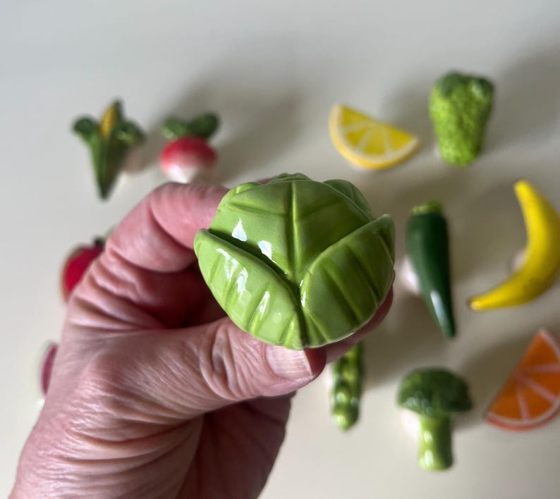 Pu&ograve; includere: Un pomello per mobili in ceramica a forma di testa di cavolo verde, tenuto in primo piano. Sullo sfondo, una collezione di altri pomelli in ceramica a forma di cibo, tra cui una fetta di limone, broccoli, banana, arancia e ravanello.