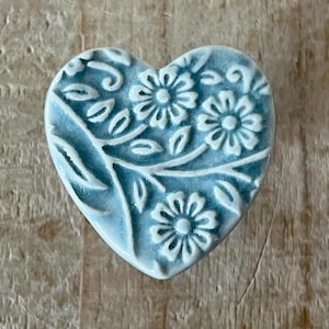 Könnte beinhalten: Ein hellblauer, herzförmiger Keramik-Knopf mit weißem Blumenmuster.