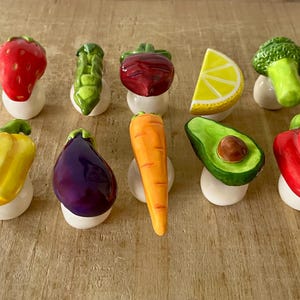 Pu&ograve; includere: Una collezione di figurine in ceramica di frutta e verdura, tra cui una fragola rossa, asparagi verdi, una barbabietola rossa, una fetta di limone giallo, un fiore di broccoli verde, un peperone giallo, una melanzana viola, una carota arancione, un avocado verde e un peperone rosso.