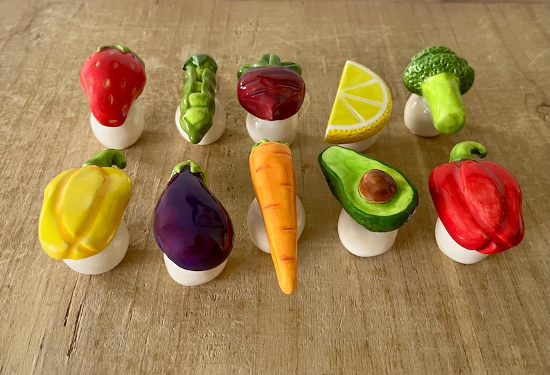 Pu&ograve; includere: Una collezione di figurine in ceramica di frutta e verdura, tra cui una fragola rossa, asparagi verdi, una barbabietola rossa, una fetta di limone giallo, un fiore di broccoli verde, un peperone giallo, una melanzana viola, una carota arancione, un avocado verde e un peperone rosso.