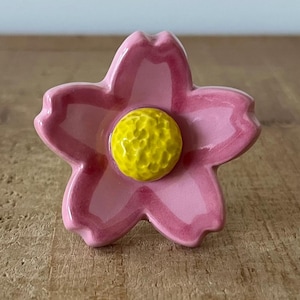 Pode incluir: Um puxador decorativo de cerâmica em forma de flor. O puxador apresenta um design rosa de cinco pétalas com um centro amarelo texturizado. O puxador de flor é provavelmente usado para móveis ou fins decorativos.