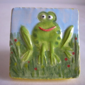 May include: A ceramic tile featuring a green frog painted on it. The frog has a red mouth and is surrounded by green grass and red flowers.