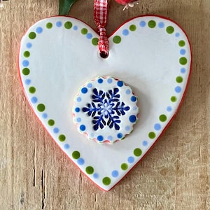 May include: White ceramic heart ornament with a red border and green and blue polka dots. A smaller white circle with a blue snowflake design is centered on the heart.